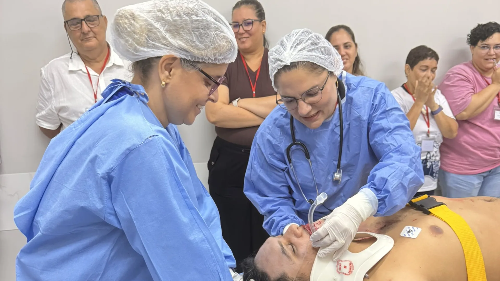 Médicas em avental cirúrgico avaliam paciente simulado em prancha rígida com colar cervical, durante o ATLS